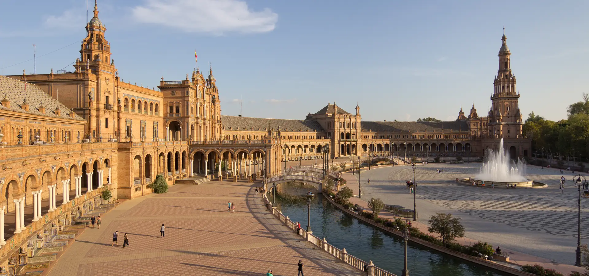 The curved brick and tiled buildings of Plaza de España in Seville, with its canal and bridges, in late-afternoon sunlight.